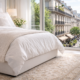 AI-generated image of a bright, elegant bedroom with white bedding, a cream throw, bedside lamp, flowers, and open French doors leading to a balcony with a Paris city view.