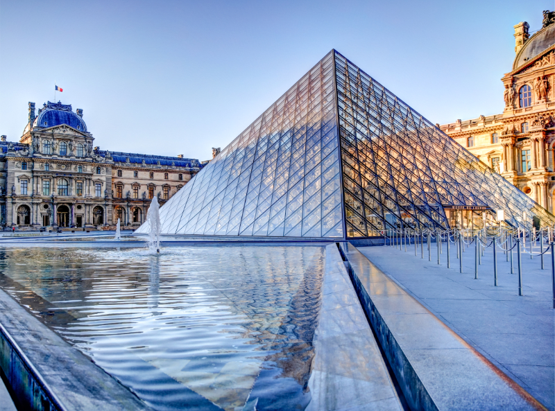 Angled view of the Louvre Pyramid in Cour Napoléon, with reflecting water, the surrounding palace façades, and the glass structure lit by daylight.