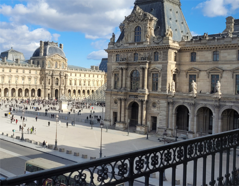 View of the Cour Napoléon at the Louvre in Paris, with the palace façades and part of the glass pyramid visible under a blue sky
