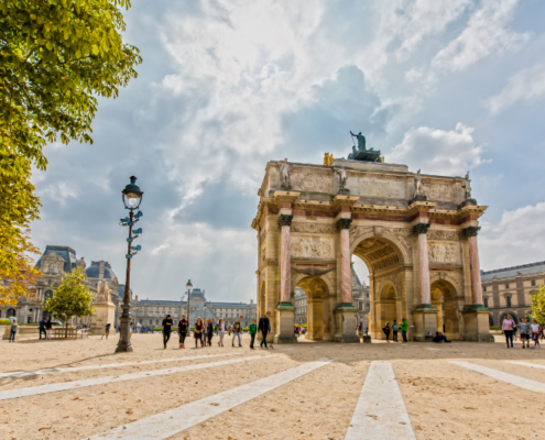 View of the Arc de Triomphe du Carrousel at the Louvre. Source: Adobe Stock View of the Arc de Triomphe du Carrousel at the Louvre in Paris, with visitors walking across the courtyard and the palace visible in the background under a dramatic sky