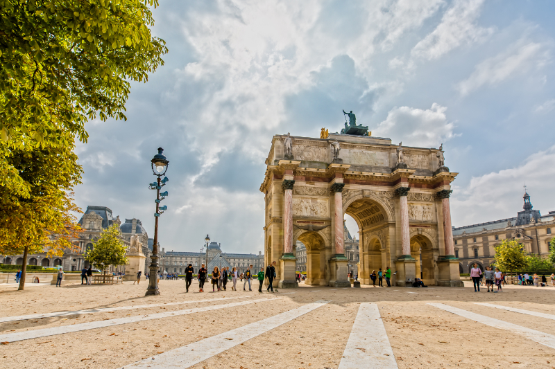 View of the Arc de Triomphe du Carrousel at the Louvre in Paris, with visitors walking across the courtyard and the palace visible in the background under a dramatic sky