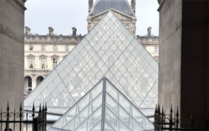 Louvre Pyramid and satellite pyramid, Cour Napoléon — I.M. Pei, 1989, viewed from the Richelieu wing arcade I.M. Pei's glass pyramid and satellite pyramid in the Cour Napoléon, viewed through a stone archway of the Louvre palace — Richelieu wing facade visible behind, iron railing in foreground, overcast Paris light