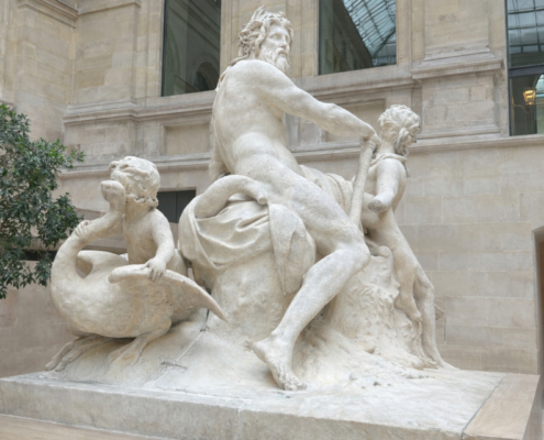 View of a monumental marble sculpture in the Cour Marly at the Louvre, shown against pale stone walls, a glass roof, and a small indoor tree, with the foreground cleared of nearby visitors.