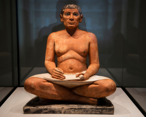View of a seated ancient Egyptian scribe statue displayed in a museum case, with painted facial features, crossed legs, and a scroll resting across the lap.