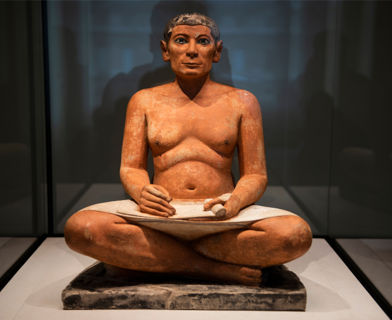 View of a seated ancient Egyptian scribe statue displayed in a museum case, with painted facial features, crossed legs, and a scroll resting across the lap.
