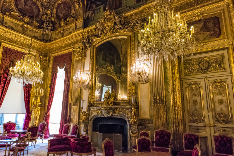 Interior view of the Apartments of Napoleon III at the Louvre in Paris, showing gilded walls, crystal chandeliers, velvet seating, and richly decorated Second Empire interiors