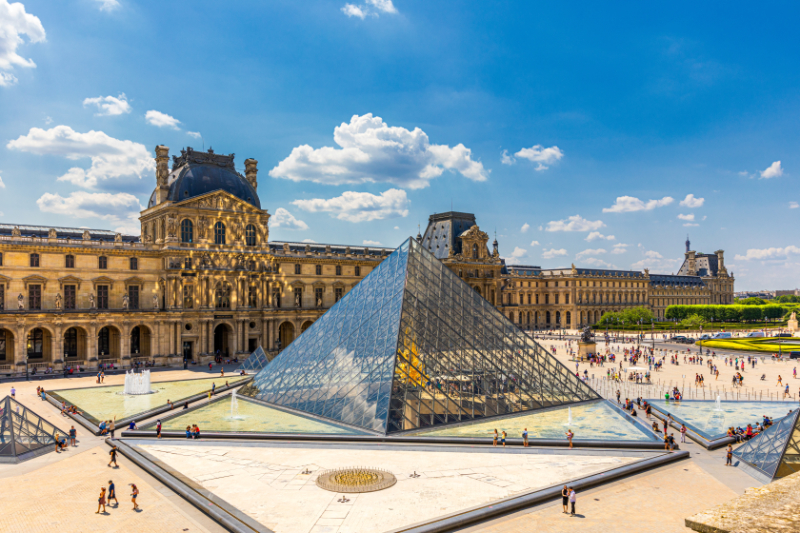 View of the Louvre Pyramid in Paris, with the surrounding palace façades and visitors crossing the Cour Napoléon under a bright blue sky