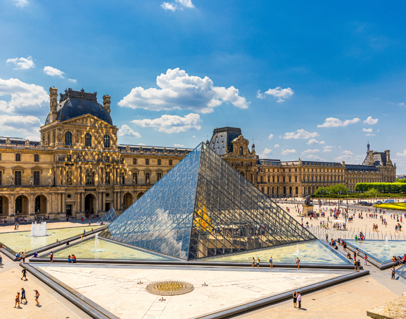 View of the Louvre Pyramid in the Cour Napoléon, with the surrounding palace façades, fountains, and visitors crossing the main courtyard.