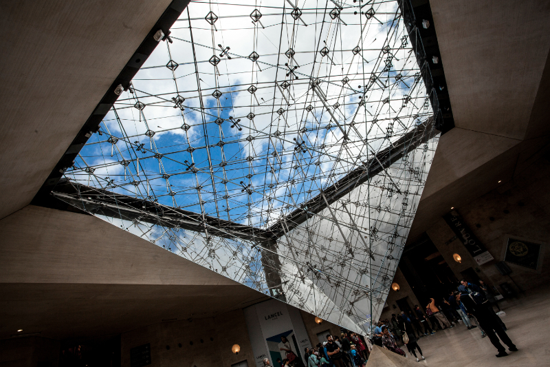 Interior view from beneath the Louvre Pyramid, showing the glass structure overhead and the central circulation space of the Hall Napoléon below.