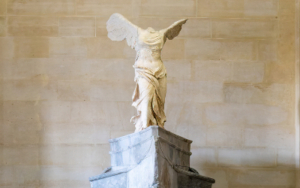 Winged Victory of Samothrace, circa 190 BCE — Daru Staircase, Denon Wing, Louvre Museum Winged Victory of Samothrace, circa 190 BCE, Louvre Museum Denon wing — low-angle view from the Daru staircase showing the marble figure on its ship-prow base, right wing spread against the vaulted stone ceiling, headless torso in wind-swept drapery