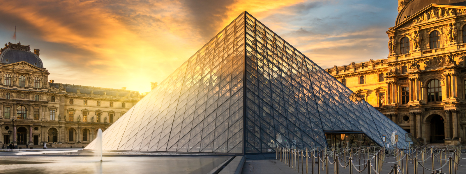 Paris Louvre Museum at sunrise. Home to Leonardo da Vinci's Mona A beautiful wide-angle shot of the glass Louvre Pyramid in Paris at sunset, with the fountain and the historic museum palace buildings in the background.