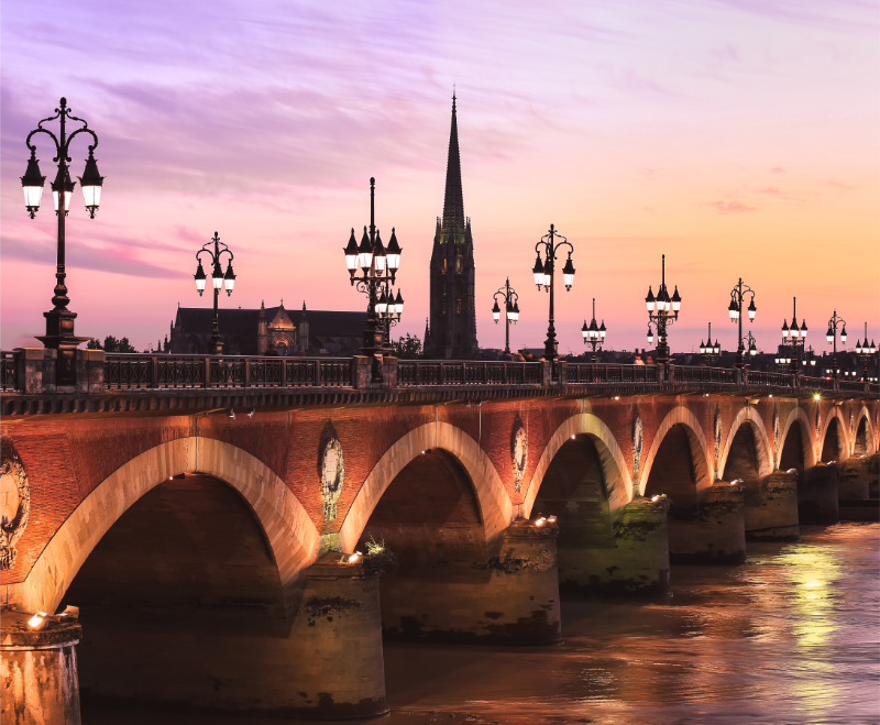 Adobe Stock image of an illuminated historic bridge over the Garonne in Bordeaux at sunset, with ornate lampposts and church spires in the background