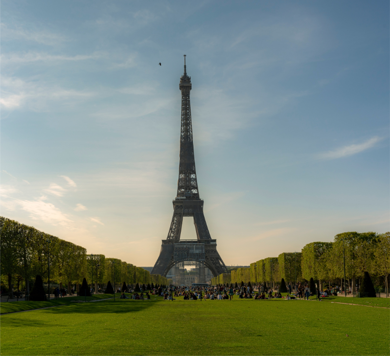 Adobe Stock photo of the Eiffel Tower viewed from the Champ de Mars in Paris, with a broad green lawn in the foreground, symmetrical rows of trimmed trees, small groups of visitors near the base, and a clear blue sky.