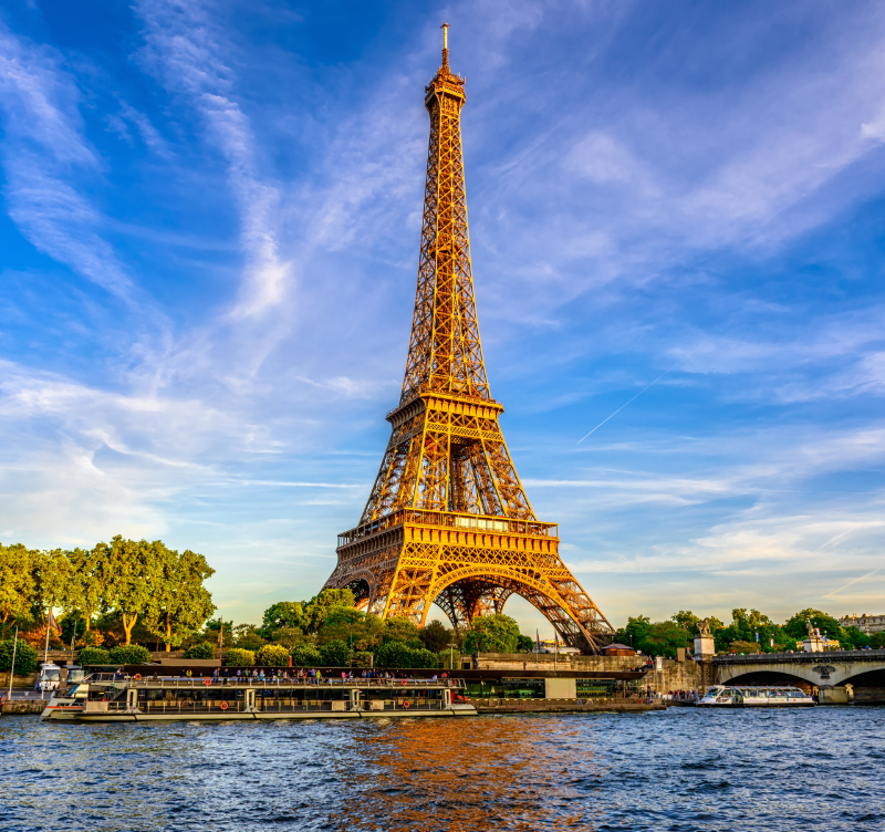 Adobe Stock photo of the Eiffel Tower seen from the Seine in Paris, with river water in the foreground, a tour boat, the bridge and embankment to the right, and a vivid blue sky with streaked white clouds.