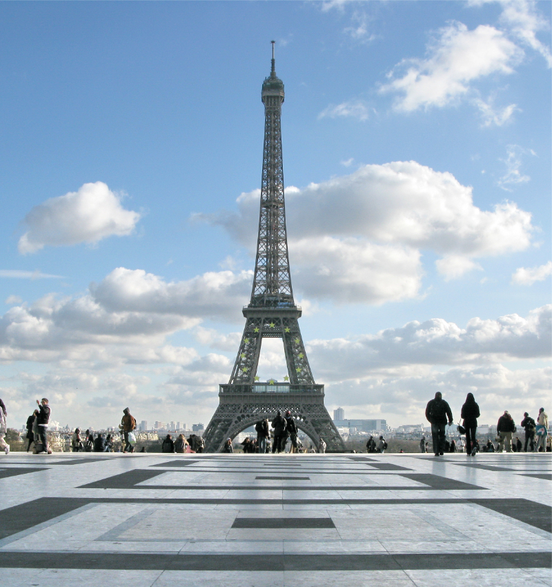 Adobe Stock photo of the Eiffel Tower seen from the Trocadéro in Paris, with the patterned stone esplanade in the foreground, scattered visitors, and a bright blue sky with large white clouds.