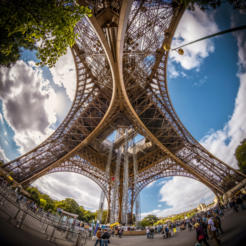 Low-angle view from beneath the Eiffel Tower, showing the sweeping curved legs, dense iron lattice, and structural geometry rising against a bright sky.