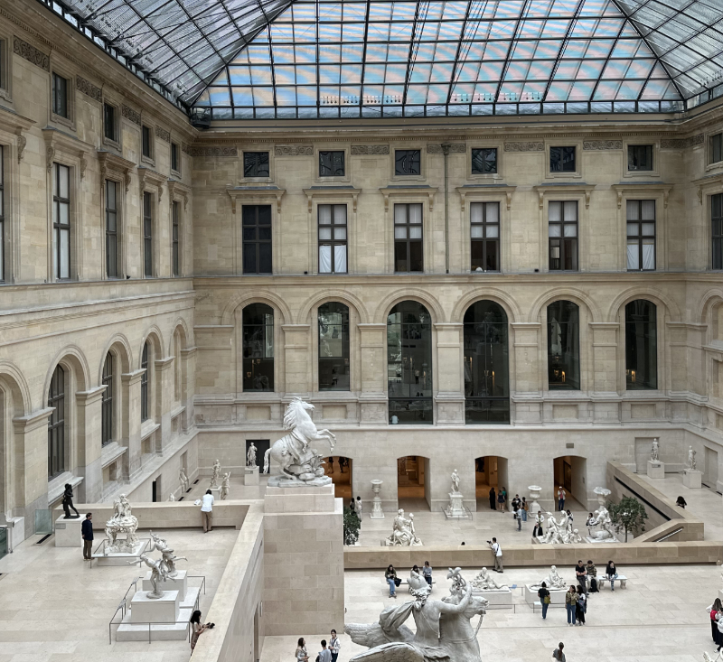 View of the Cour Marly at the Louvre, showing the skylit sculpture courtyard with pale stone façades, monumental marble statues, and visitors moving through the open interior space.