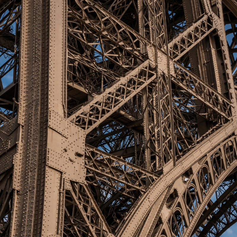 AI-generated square close-up of the Eiffel Tower’s dense iron structure, showing rivets, intersecting beams, lattice trusses, and precise metal assemblies in warm daylight.