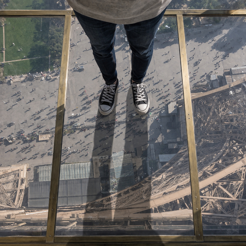 AI-generated square top-down view of a single person standing on the Eiffel Tower’s glass floor, with straight metal bars framing the transparent panels and the esplanade visible far below.