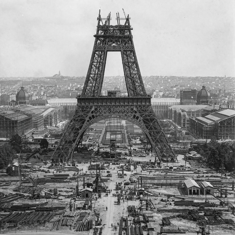 AI-generated square black-and-white historical-style view of the Eiffel Tower during construction, showing the lower structure rising above a busy worksite with exhibition buildings and the Paris skyline in the background.
