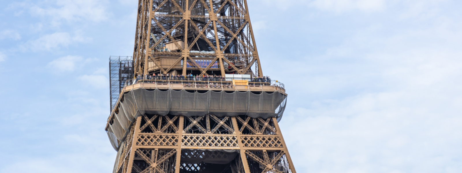 Exterior view of the first floor of the Eiffel Tower, showing visitors along the platform, the iron lattice structure above, and protective netting installed beneath the deck against a cloudy sky.