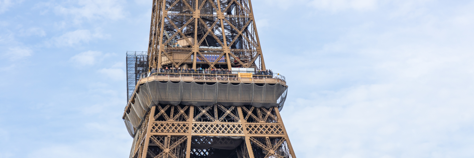Second floor of the Eiffel Tower on sunny day with protective ne Exterior view of the first floor of the Eiffel Tower, showing visitors along the platform, the iron lattice structure above, and protective netting installed beneath the deck against a cloudy sky.