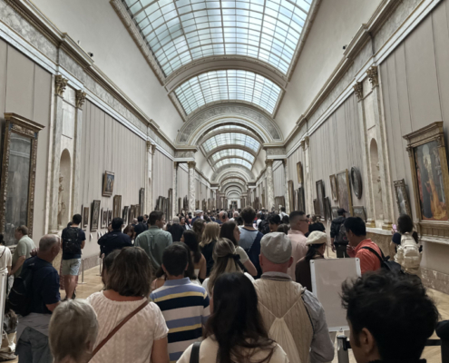 View of La Grande Galerie at the Louvre with visitors. View of La Grande Galerie at the Louvre, showing the long skylit corridor lined with paintings and crowded with visitors moving through the gallery.