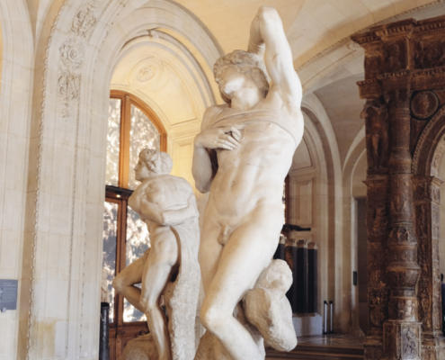 Adobe Stock view of marble sculptures at the Louvre. View of two marble nude sculptures displayed in an ornate gallery at the Louvre, with vaulted ceilings, carved stone arches, and patterned marble floors.