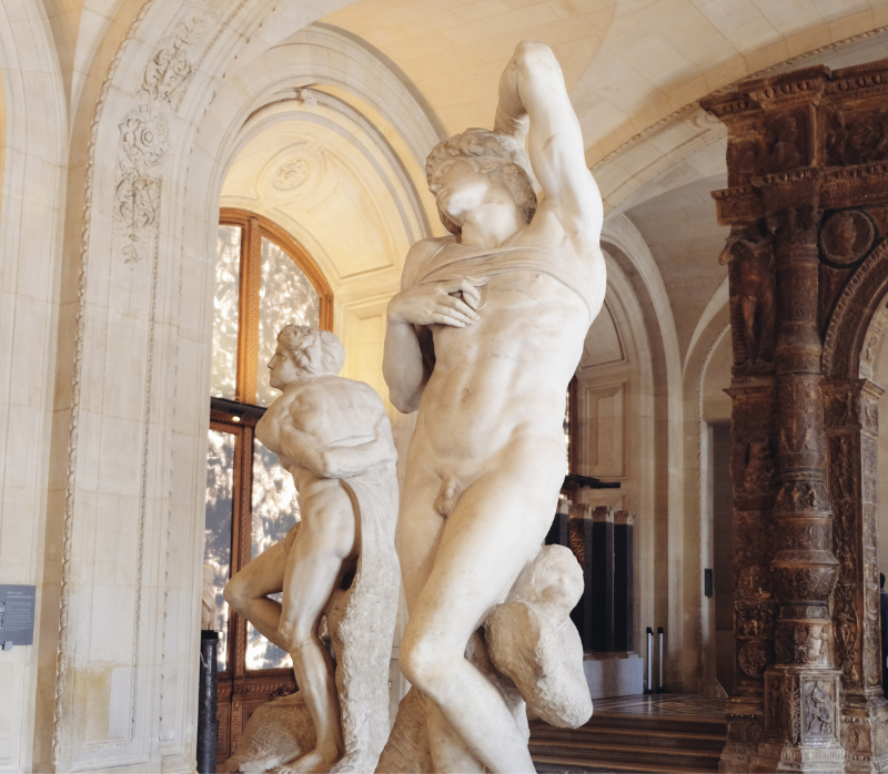 View of two marble nude sculptures displayed in an ornate gallery at the Louvre, with vaulted ceilings, carved stone arches, and patterned marble floors.