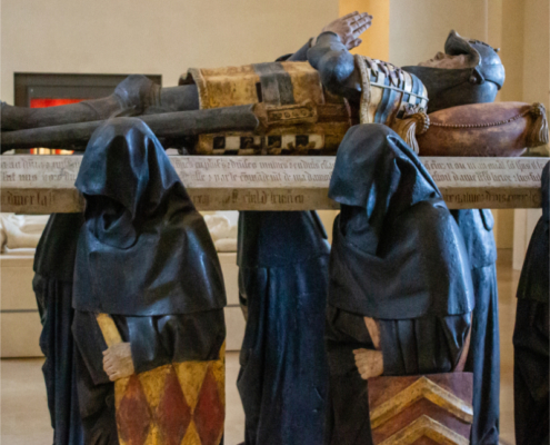 Adobe Stock view of a medieval funerary sculpture at the Louvre. View of a medieval funerary monument at the Louvre, showing a recumbent armored figure resting on a sculpted tomb supported by hooded mourners in a museum gallery.