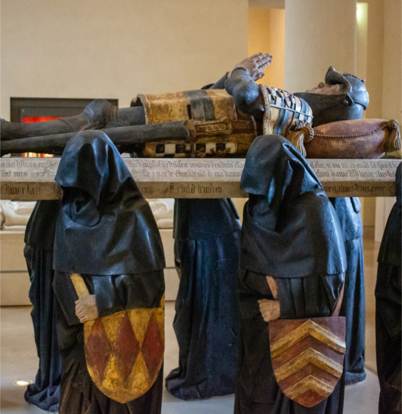 View of a medieval funerary monument at the Louvre, showing a recumbent armored figure resting on a sculpted tomb supported by hooded mourners in a museum gallery.