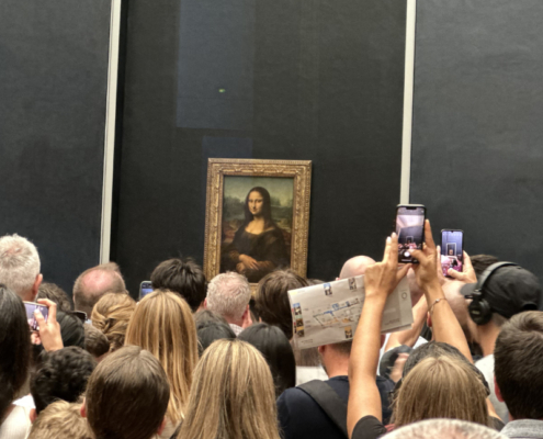 View of the Mona Lisa at the Louvre surrounded by visitors. View of the Mona Lisa at the Louvre, showing the painting displayed behind protective glass with a large crowd of visitors gathered in front of it.