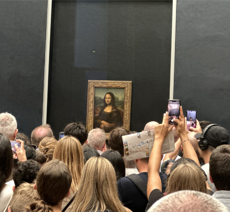View of the Mona Lisa at the Louvre, showing the painting displayed behind protective glass with a large crowd of visitors gathered in front of it.