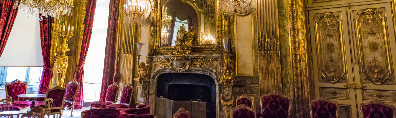 The Apartments of Napoleon III at the Louvre - Adobe Stock View of a richly decorated salon in the Apartments of Napoleon III at the Louvre, with gilded walls, crystal chandeliers, red velvet seating, and an ornate fireplace beneath a large mirror.