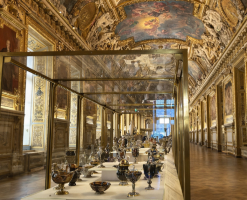 Edited view of the Galerie d’Apollon at the Louvre. Edited view of the Galerie d’Apollon at the Louvre, showing the ornate gilded ceiling, painted vaults, framed portraits, and central display cases filled with decorative objects, with the foreground cleared of nearby visitors.