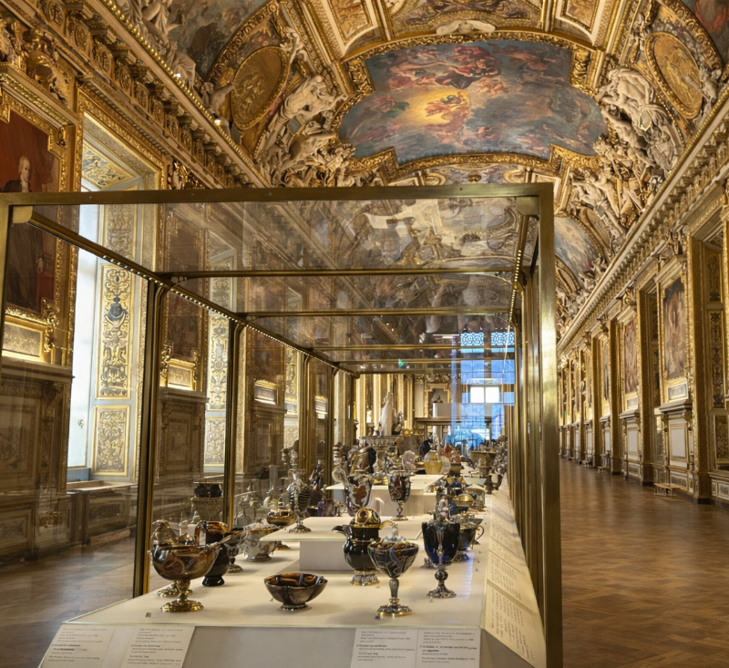 Edited view of the Galerie d’Apollon at the Louvre, showing the ornate gilded ceiling, painted vaults, framed portraits, and central display cases filled with decorative objects, with the foreground cleared of nearby visitors.