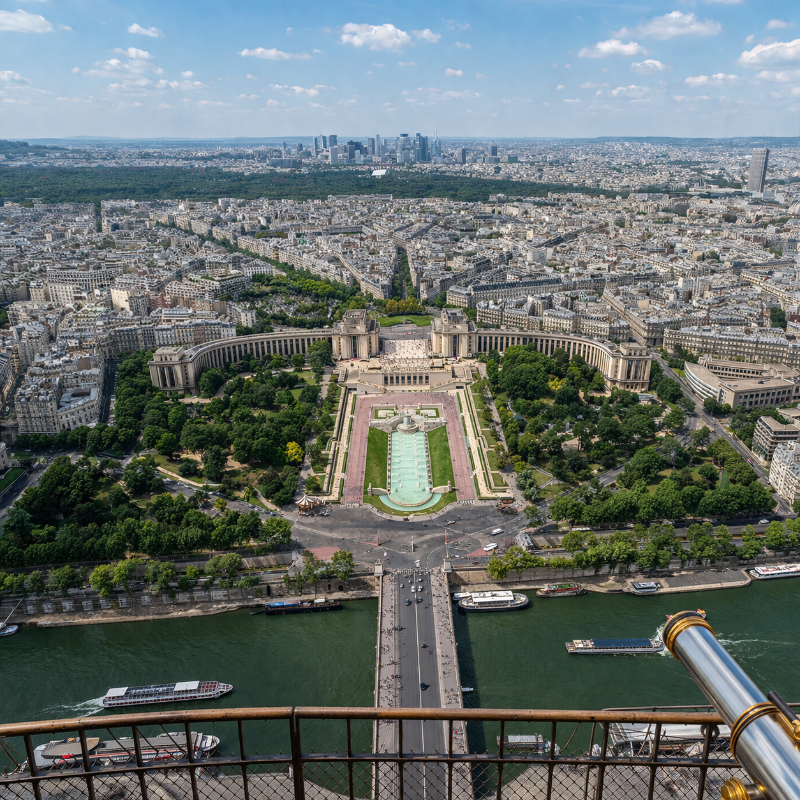 AI-generated panoramic view from the Eiffel Tower looking over the Trocadéro gardens and the Palais de Chaillot, with La Défense visible in the distance under clear daylight.
